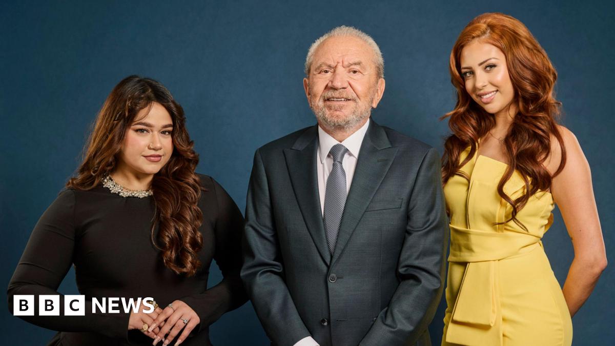 Three people pose for a studio photograph against a dark blue backdrop. Lord Alan Sugar stands in the centre wearing a dark suit and tie. Karishma Vijay stands to one side wearing a black dress, while finalist Pascha Myhill stands on the other side wearing a yellow sleeveless outfit.