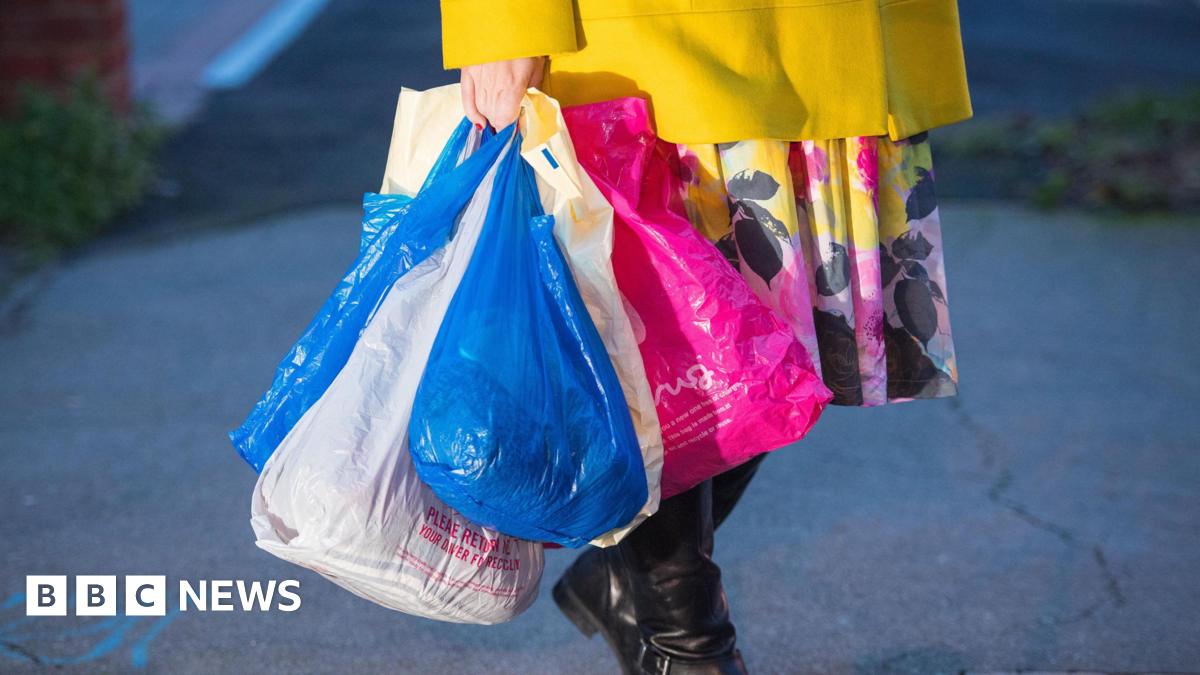 A woman carries shopping in plastic carrier bags. The woman is holding about five bags in one hand. She is wearing a floral skirt with a yellow jacket.
