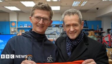 Alexander Armstrong is on the right and has grey hair with a black coat and blue and white scarf. He is holding one side of a red top that reads "one big thank you" in white writing. On the left is Michael who has brown hair and glasses and is wearing a blue navy hoodie, and is holding the other side of the top.