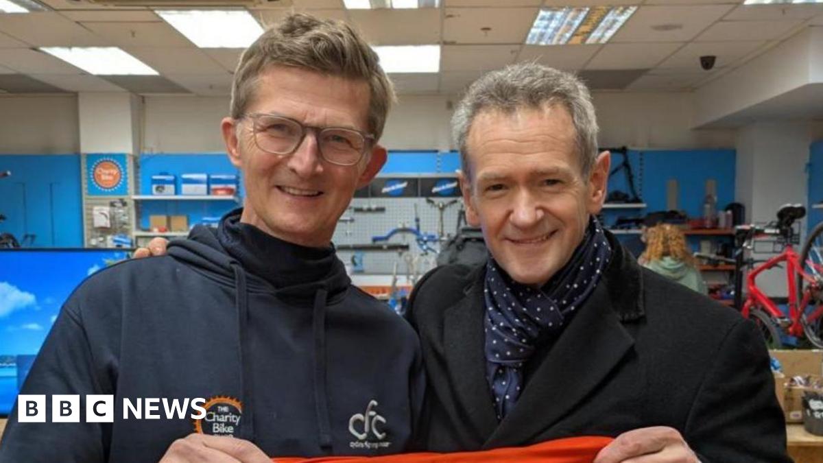Alexander Armstrong is on the right and has grey hair with a black coat and blue and white scarf. He is holding one side of a red top that reads "one big thank you" in white writing. On the left is Michael who has brown hair and glasses and is wearing a blue navy hoodie, and is holding the other side of the top.