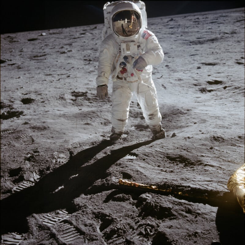 An astronaut in a white spacesuit stands on the gray, dusty surface of the Moon, with boot prints and equipment visible. The reflection of another astronaut and the lunar module is seen in the helmet visor.