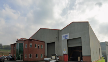A large grey warehouse with red brick offices built on the front of it is pictured on a cloudy day. A flat bed truck is parked outside the main shutters to the warehouse.