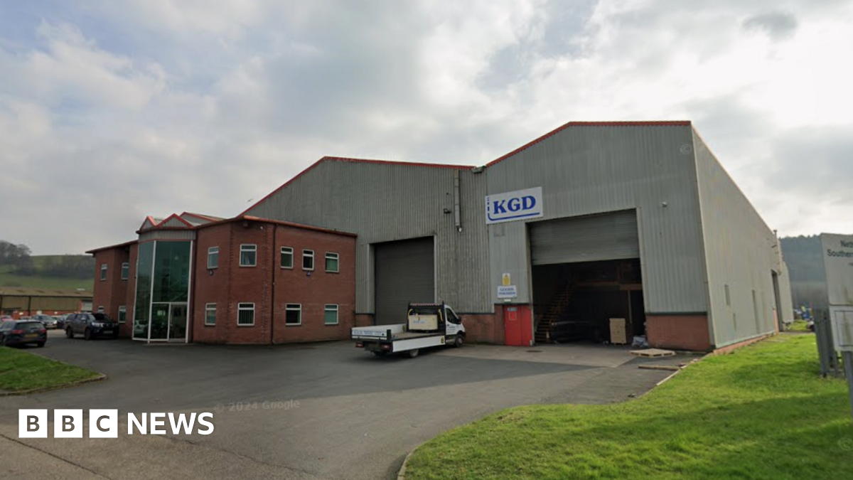 A large grey warehouse with red brick offices built on the front of it is pictured on a cloudy day. A flat bed truck is parked outside the main shutters to the warehouse.