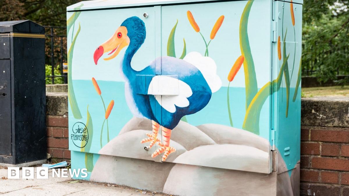A mural of a blue dodo painted on to a signal box in Shrewsbury. The dodo is standing on a grey rock surrounded by green reeds. In the background is blue water.
