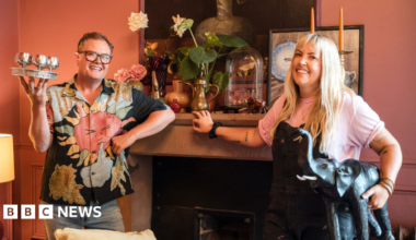 Alan Carr holding silver wine glasses and a woman holding an elephant statue in front of a fire place
