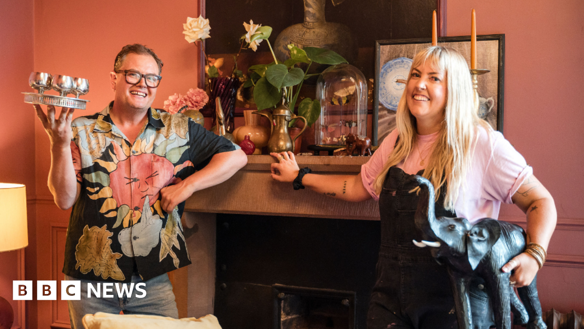 Alan Carr holding silver wine glasses and a woman holding an elephant statue in front of a fire place