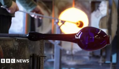 Photo inside Bristol's blue glass factory. This picture shows a blue vase being shaped on a stick in the factory. In the background you can see a man putting a a long stick into furnace.