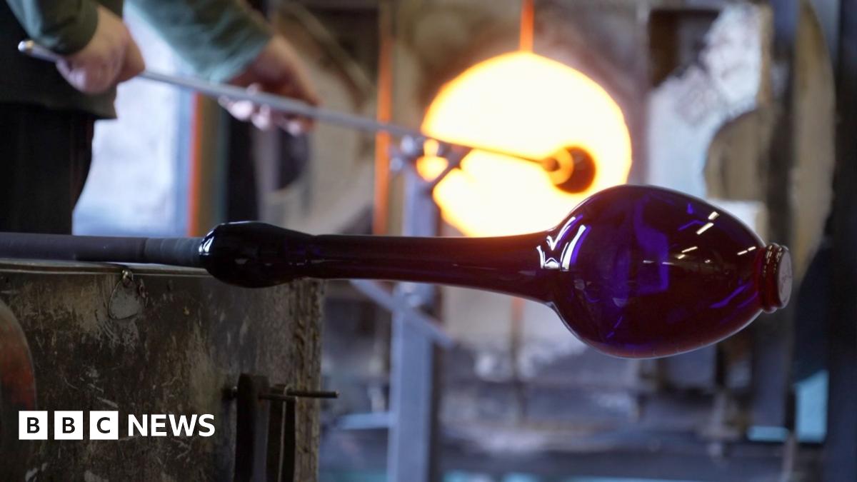 Photo inside Bristol's blue glass factory. This picture shows a blue vase being shaped on a stick in the factory. In the background you can see a man putting a a long stick into furnace.