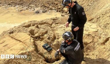 Two bomb disposal experts stand over a WW2 bomb that has been rigged with explosives, surrounded by sand.