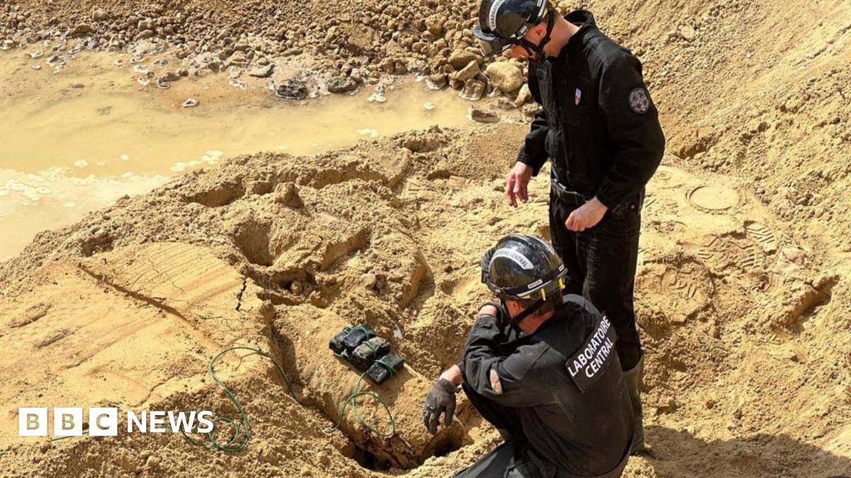 Two bomb disposal experts stand over a WW2 bomb that has been rigged with explosives, surrounded by sand.