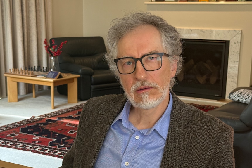A man with glasses and a grey jacket, sits inside a stylish lounge room. 