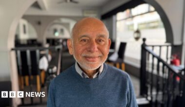 Nathan sitting in a pub with a drink. He is wearing a blue tracksuit top and has his arms crossed. He has a short beard and dark brown hair.