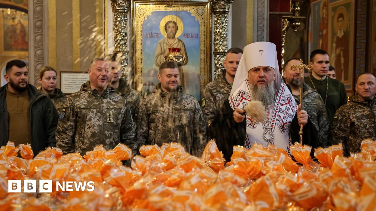 Metropolitan Epiphanius I, head of the Orthodox Church of Ukraine, sprinkles holy water on packets holding Easter cakes. Ukrainian service men stand behind him.