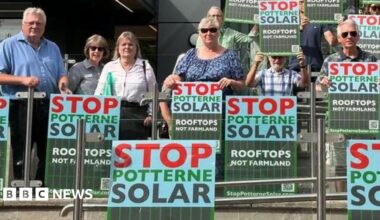 A group of residents holding up "stop potterne solar - rooftops not farmland" signs. They are standing against a glass barrier with a grey office building in the background.