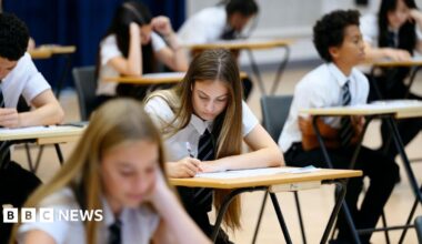 Teenage girl wearing school shirt and tie writing in exam booklet, arm resting on desk