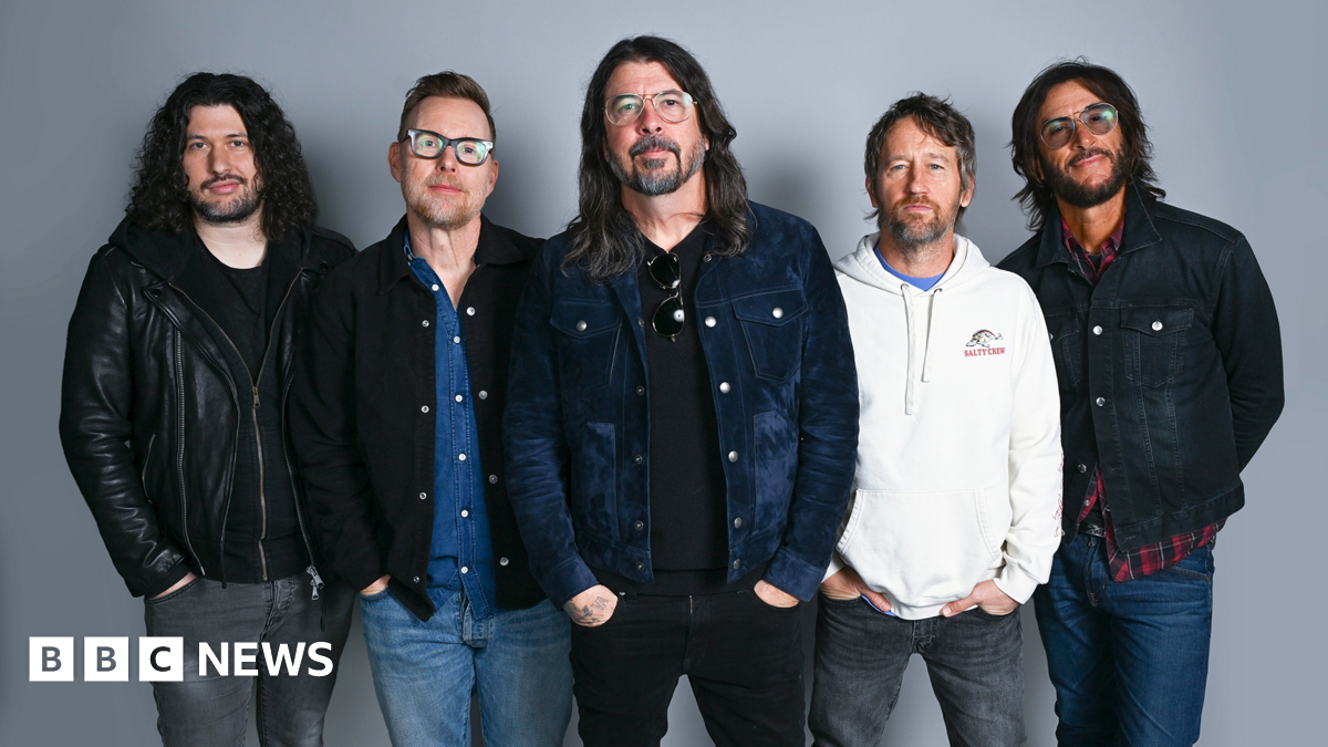 Five members of the Foo Fighters band stand side by side looking directly at the camera, hands in pockets.