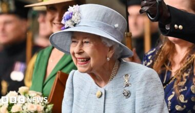 King Charles III with his late mother, Queen Elizabeth II - both are smiling off camera, and standing on the Buckingham Palace balcony which can just be seen. The late Queen wears an emerald green hat and jacket, with pearls and a diamond brooch in the shape of a bow, while the King has a navy pin-striped suit and a pale blue patterned tie