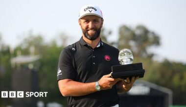 Jon Rahm, with a dark beard and wearing a black polo shirt, smiles as he holds the LIV Mexico trophy - a black base with a glass skull