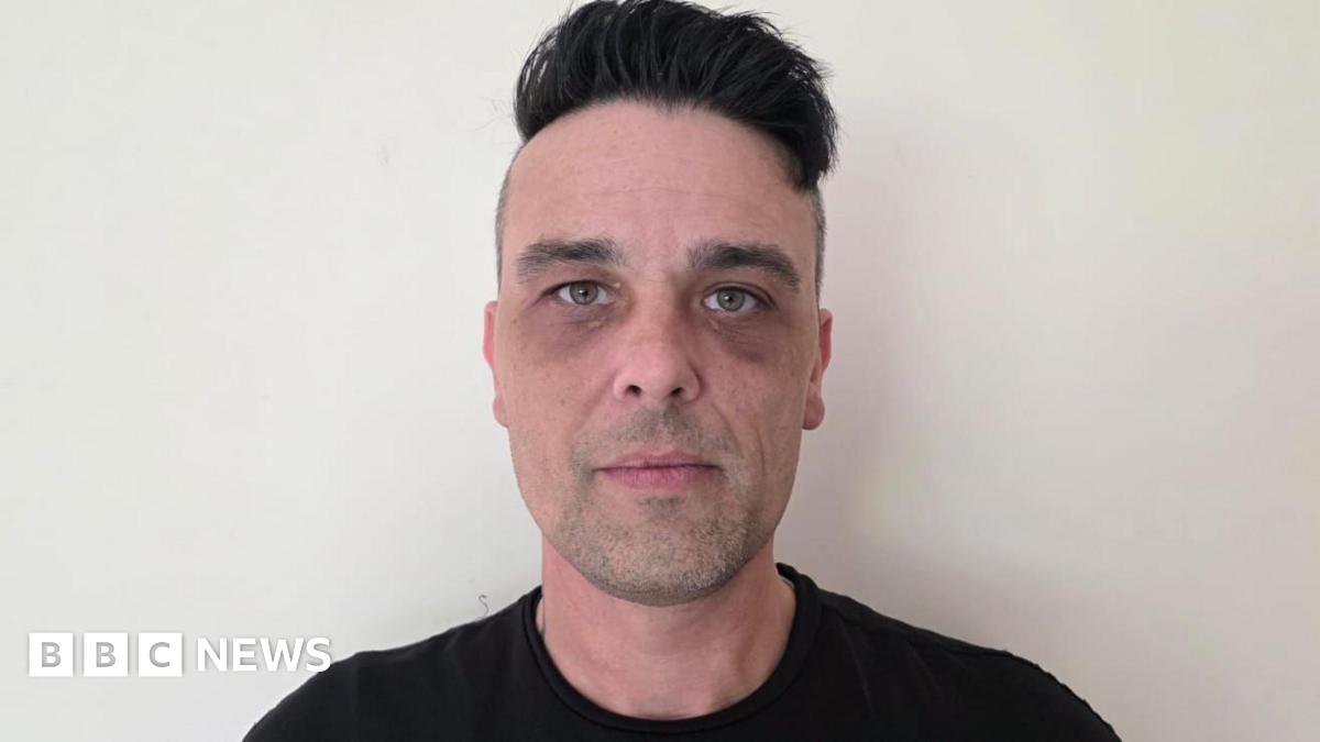 Kevin Brewer has black hair. He's wearing a black top and standing against a white wall.