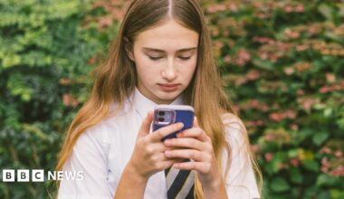 A teenage girl with long blonde hair wearing a school shirt and tie is looking at her mobile phone, which has a blue cover.