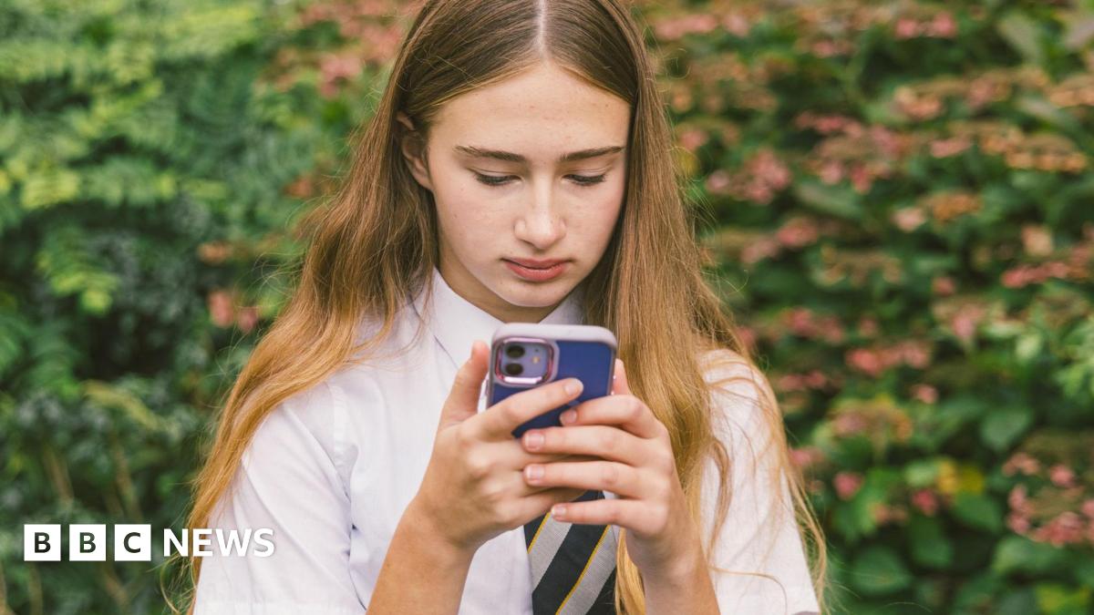 A teenage girl with long blonde hair wearing a school shirt and tie is looking at her mobile phone, which has a blue cover.