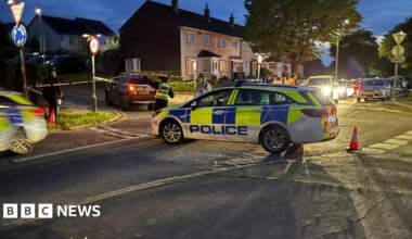 Two police cars on a road. There is police tape on sign posts. There is a car driving outside of the cordon and a person in police uniform directing them. There are houses in the background and cars with their headlights on.