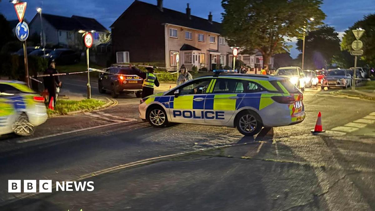 Two police cars on a road. There is police tape on sign posts. There is a car driving outside of the cordon and a person in police uniform directing them. There are houses in the background and cars with their headlights on.