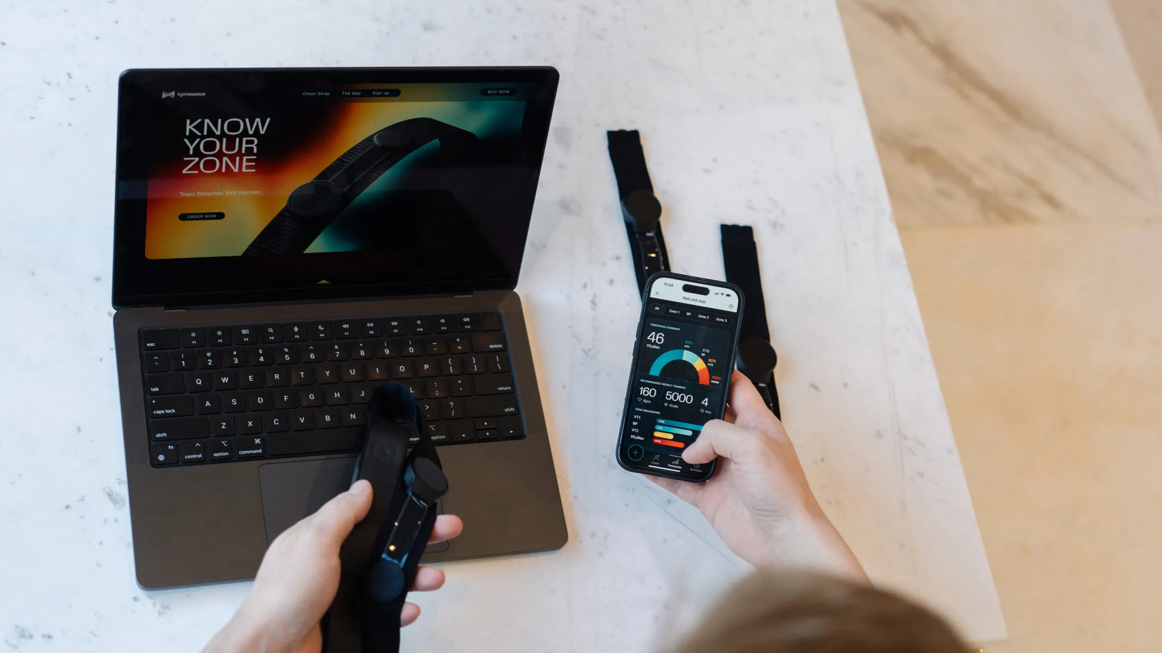A laptop displaying the Tymewear website, a smartphone showing the Tymewear app, and Tymewear chest straps.