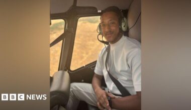A man in a white shirt sits in a helicopter with a headset on. He smiles at the camera.