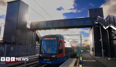 A red and blue Supertram is driving on the tracks under a metal pedestrian bridge
