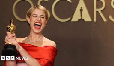Jessie Buckley with her eyes closed smiling widely at the camera. She is holding an Oscar award and is wearing a red and pink satin dress.