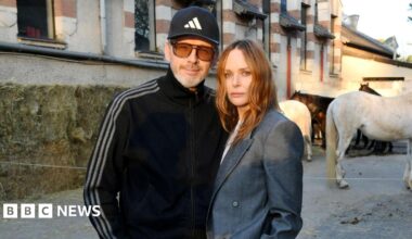 Alasdhair Willis is wearing a dark blue cap and tracksuit top and a pair of sunglasses with brown lenses. Stella McCartney has shoulder length brown hair and is wearing a grey suit jacket. The couple are standing outside a large building and there are four horses in the background.