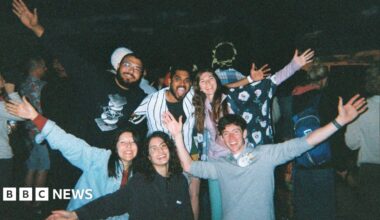 A group of young people. Some are standing and some are kneeling and they all have their arms outstretched. They are all smiling widely at the camera. There are three young women and three young men and they appear to be at a festival. It is dark behind them and other people can be seen in the background.