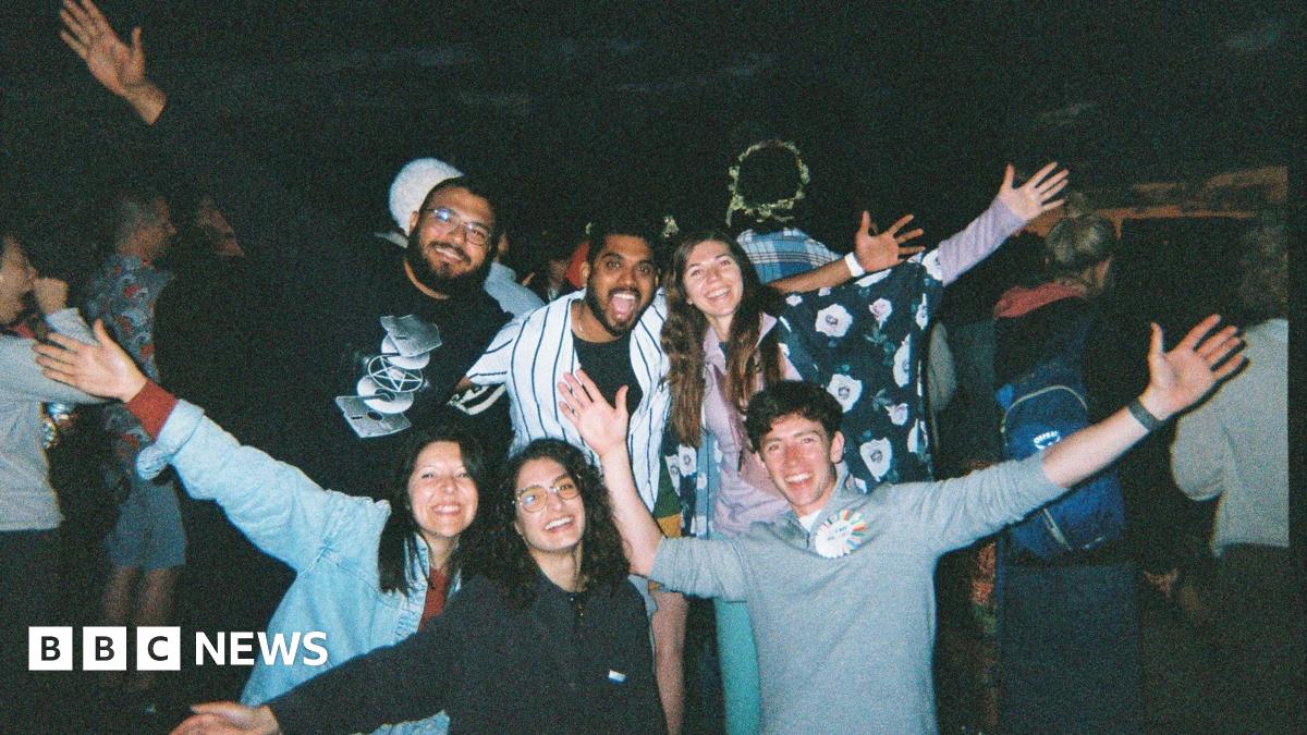 A group of young people. Some are standing and some are kneeling and they all have their arms outstretched. They are all smiling widely at the camera. There are three young women and three young men and they appear to be at a festival. It is dark behind them and other people can be seen in the background.