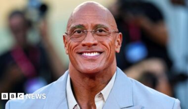 Dwayne Johnson smiles at the camera at The Smashing Machine red carpet event during last year's Venice International Film Festival. He is wearing an open-necked white shirt and a powder blue suit jacket.
