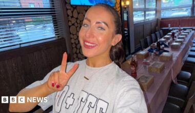 A selfie of Ashlin McCourt wearing a grey jersey sweatshirt with her dark hair slicked back. She is smiling and making a peace sign while working as a waitress with a long table seen behind her