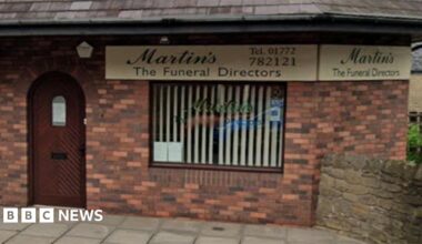 Streetview image of the funeral directors in Longridge. A red brick building with a brown arched door to the left and a window to the right with a vertical cream blind.