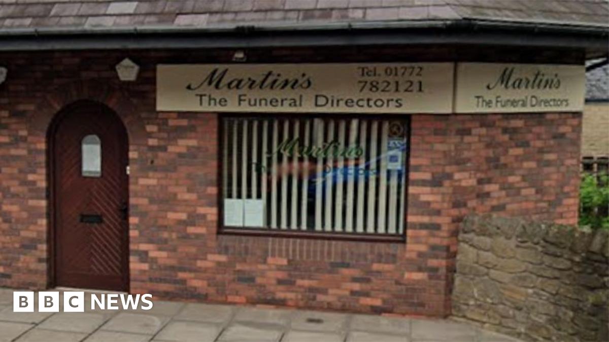 Streetview image of the funeral directors in Longridge. A red brick building with a brown arched door to the left and a window to the right with a vertical cream blind.