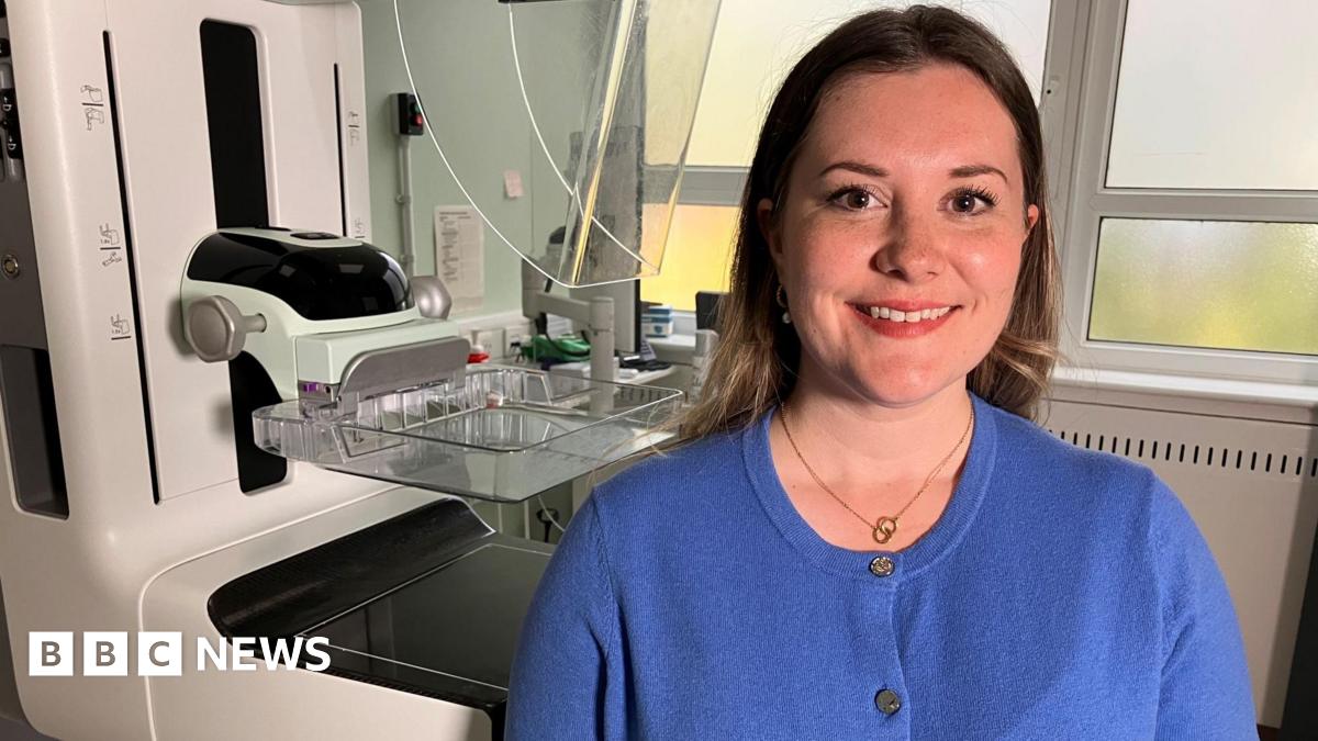 Rebecca Corbett, Quality and Improvement Manager for West Sussex Brighton and Hove Breast screening Service. Rebecca, who is sat in front of the screening machine, is wearing a blue t-shirt and has her hair down.