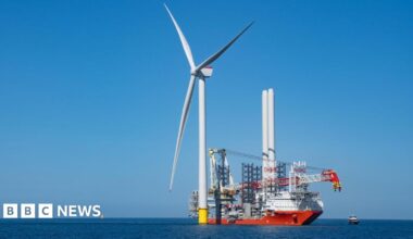 A wind turbine in the sea. Next to it, there is a large ship being used in the turbine construction process. The blue sea is flat and calm. The cloudless sky is blue.