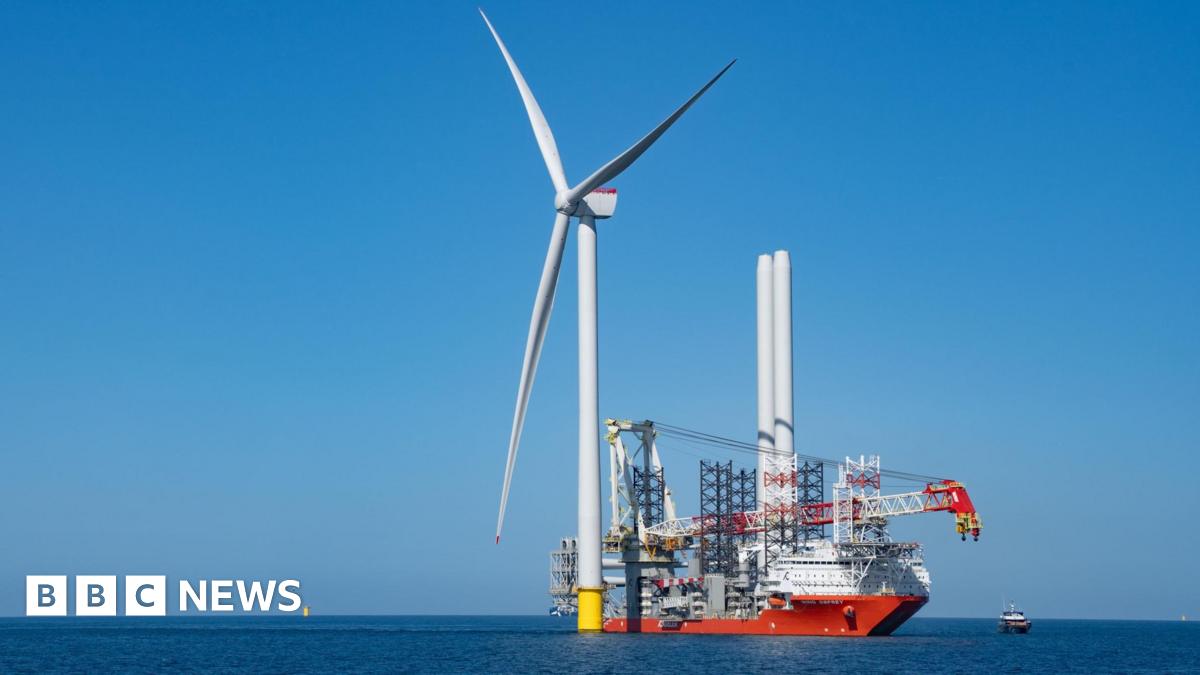 A wind turbine in the sea. Next to it, there is a large ship being used in the turbine construction process. The blue sea is flat and calm. The cloudless sky is blue.