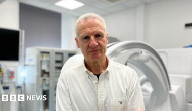 Tom Binet, a man with short white hair, is wearing a white shirt and no tie. He is standing in front of medical equipment, in a room which appears to be in a hospital. He is staring directly at the camera.
