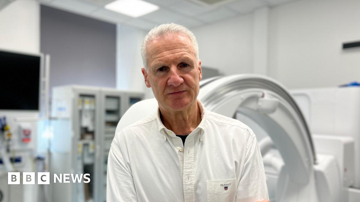 Tom Binet, a man with short white hair, is wearing a white shirt and no tie. He is standing in front of medical equipment, in a room which appears to be in a hospital. He is staring directly at the camera.
