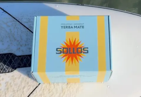 Box of Sollos Pineapple & Coconut Yerba Mate on a surfboard in clear water.