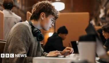 A person with short curly hair types on a laptop at a wooden desk in a library and study space, wearing glasses and headphones, with other people working nearby in the background.