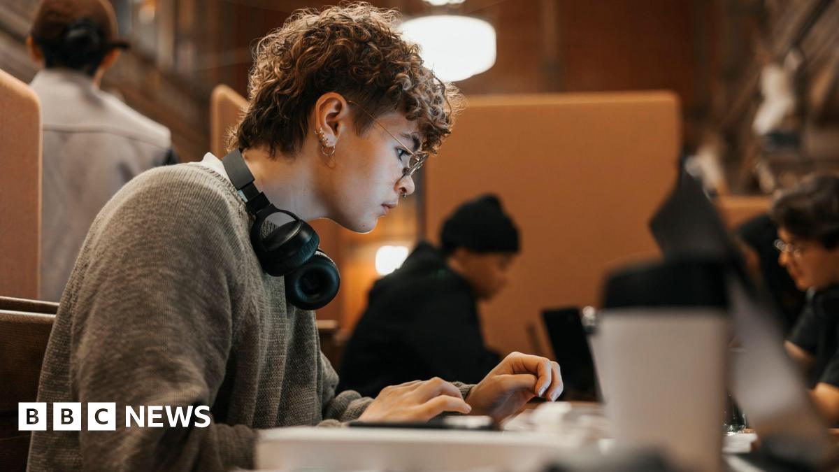 A person with short curly hair types on a laptop at a wooden desk in a library and study space, wearing glasses and headphones, with other people working nearby in the background.