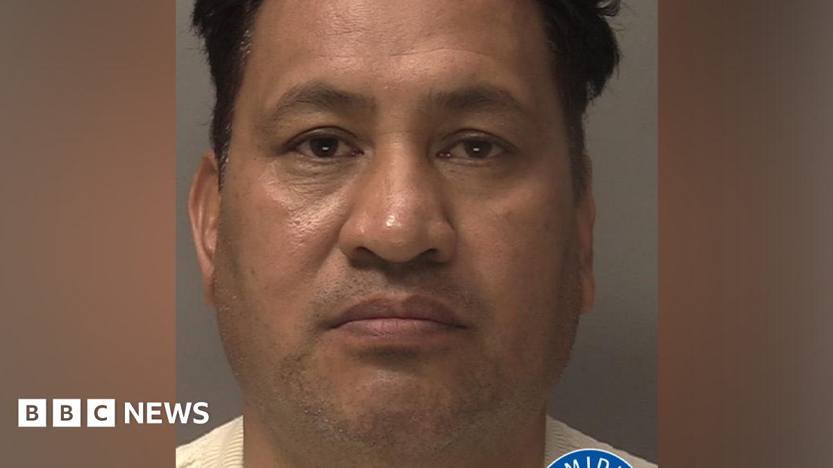 A police mugshot of a man with dark hair. He is wearing a cream jumper. There is a West Midlands Police logo in the bottom right corner.