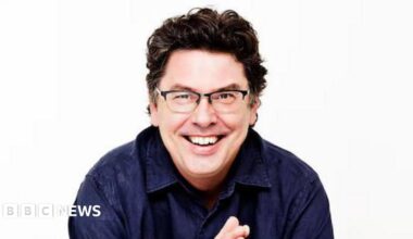A man with brown curly hair, wearing glasses and a dark blue shirt, smiling at the camera