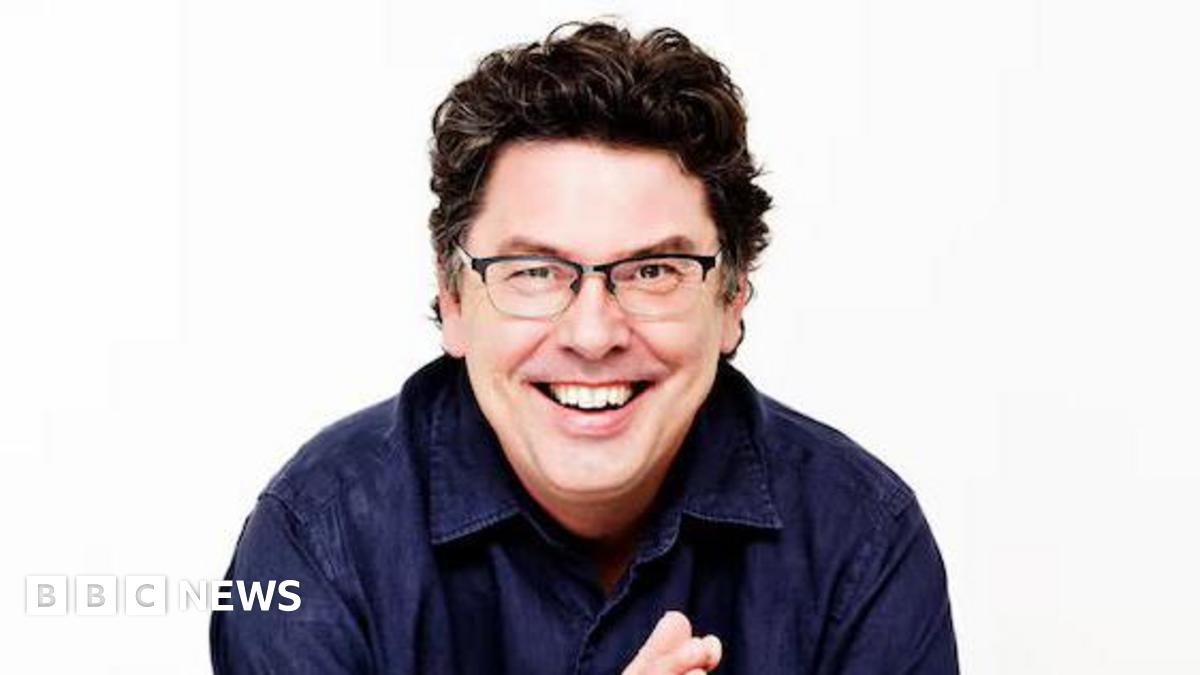 A man with brown curly hair, wearing glasses and a dark blue shirt, smiling at the camera