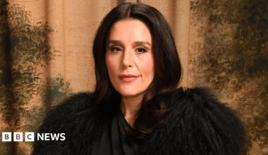 Jessie Ware poses for a photographer. She has a slight smile on her face. She is wearing a dramatic black outfit, covering her shoulders with feathery fabric. She has long, dark hair and is in front of a patterned backdrop of earthy colours.